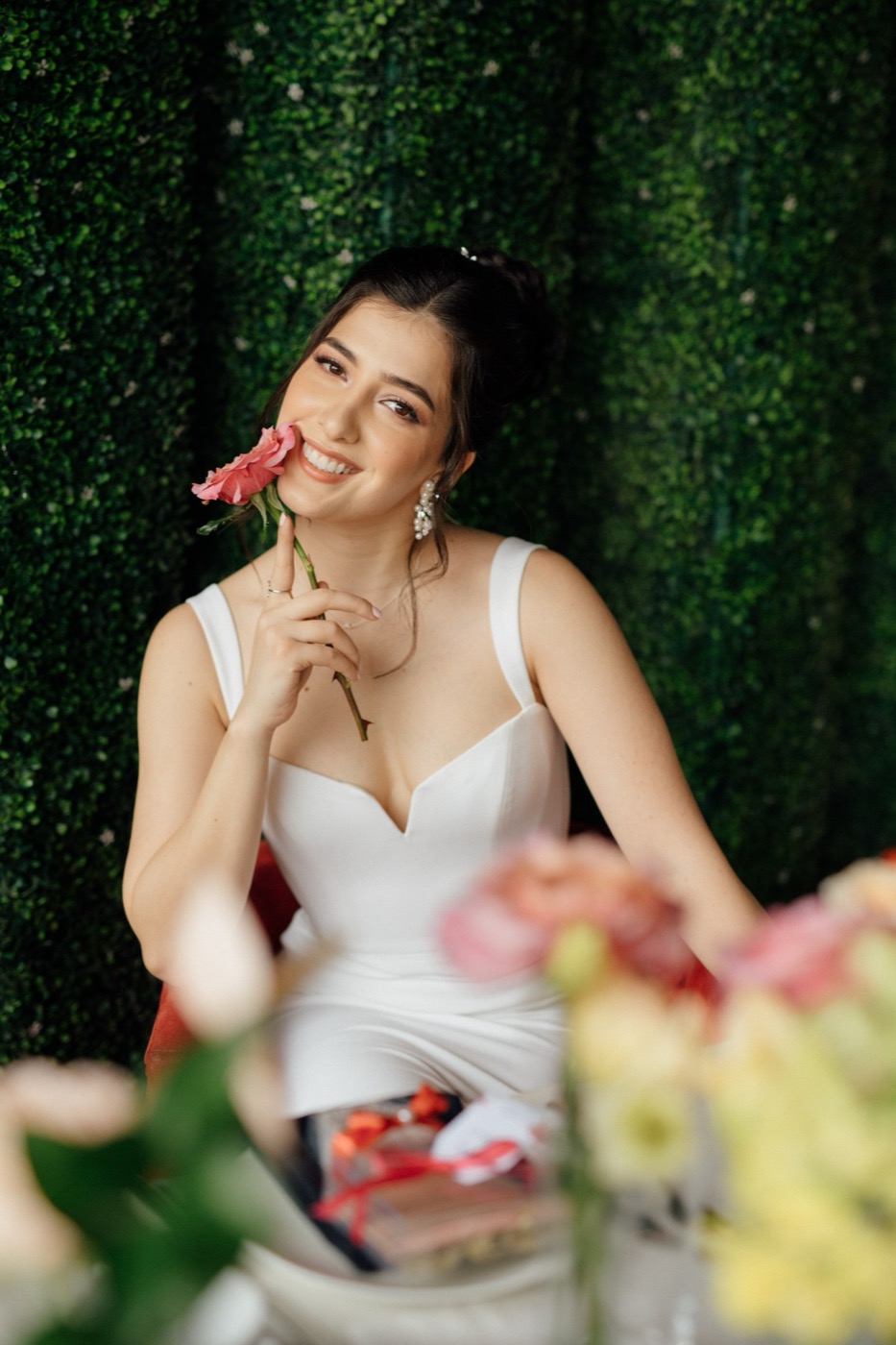 Abril smiling, holding a pink rose to her cheek in front of a green hedge wall